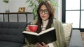 Young beautiful hispanic woman reading book and drinking coffee sitting on sofa at home Royalty Free Stock Photo