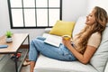 Young beautiful hispanic woman reading book and drinking coffee sitting on sofa at home Royalty Free Stock Photo