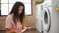Young beautiful hispanic woman reading washing machine instruction at laundry room