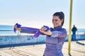 Young beautiful hispanic woman stretching arms using elastic band at seaside Royalty Free Stock Photo