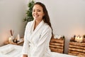 Young beautiful hispanic woman wearing bathrobe sitting on massage board at beauty salon