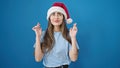 Young beautiful hispanic woman wearing christmas hat crossing fingers for luck over isolated blue background