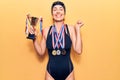 Young beautiful hispanic woman wearing swimmer swimwear and winner medals holding trophy screaming proud, celebrating victory and Royalty Free Stock Photo