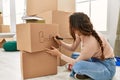 Young beautiful hispanic woman writing books on cardboard box sitting on floor at new home Royalty Free Stock Photo