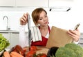 Young beautiful home cook woman in red apron at domestic kitchen reading cookbook following recipe holding knife Royalty Free Stock Photo