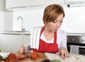 Young beautiful home cook woman in red apron at domestic kitchen reading cookbook following recipe holding knife