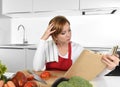 Young beautiful home cook woman in red apron at modern domestic kitchen reading cookbook following recipe Royalty Free Stock Photo