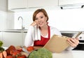 Young beautiful home cook woman in red apron at modern domestic kitchen reading cookbook following recipe Royalty Free Stock Photo