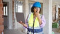 Young beautiful latin woman builder using laptop talking by walkie talkie at construction site Royalty Free Stock Photo