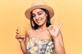 Young beautiful latin woman wearing summer hat drinking glass of orange juice doing ok sign with fingers, smiling friendly Royalty Free Stock Photo