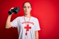 Young beautiful lifeguard woman wearing t-shirt with red cross and whistle using binoculars with a confident expression on smart