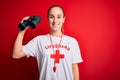 Young beautiful lifeguard woman wearing t-shirt with red cross and whistle using binoculars with a happy face standing and smiling