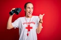 Young beautiful lifeguard woman wearing t-shirt with red cross and whistle using binoculars pointing and showing with thumb up to