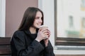 Young beautiful millenial girl in a coat is sitting on an outdor bench with a paper cup of coffee. Autumn or spring day Royalty Free Stock Photo