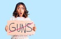 Young beautiful mixed race woman holding no guns warning banner scared and amazed with open mouth for surprise, disbelief face