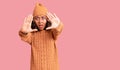 Young beautiful mixed race woman wearing wool sweater and winter hat doing frame using hands palms and fingers, camera perspective Royalty Free Stock Photo