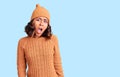 Young beautiful mixed race woman wearing wool sweater and winter hat in shock face, looking skeptical and sarcastic, surprised