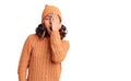 Young beautiful mixed race woman wearing wool sweater and winter hat yawning tired covering half face, eye and mouth with hand Royalty Free Stock Photo