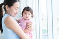 Young beautiful mother holding and looking at her cute little Caucasian 7 months newborn baby on her hands, standing near window