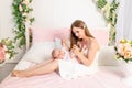 A young beautiful mother holds her daughter a girl of 6 months on her lap on a white bed, playing and kissing her, place for text Royalty Free Stock Photo