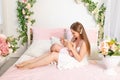 A young beautiful mother holds her daughter a girl of 6 months on her lap on a white bed, playing and kissing her, place for text Royalty Free Stock Photo