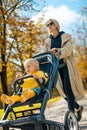 Young beautiful mother wearing a rain coat pushing stroller with her little baby boy child, walking in city park on a Royalty Free Stock Photo