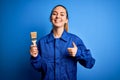 Young beautiful painter woman with blue eyes painting wearing uniform using paint brush happy with big smile doing ok sign, thumb Royalty Free Stock Photo