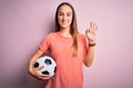 Young beautiful player woman playing soccer holding football ball  over pink background doing ok sign with fingers, excellent Royalty Free Stock Photo