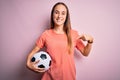 Young beautiful player woman playing soccer holding football ball  over pink background with surprise face pointing finger to Royalty Free Stock Photo