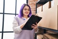 Young beautiful plus size woman ecommerce business worker writing on document standing by shelving at office