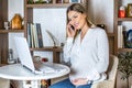 Young beautiful pregnant girl talking on her phone while working on her computer Royalty Free Stock Photo