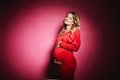 Young beautiful pregnant woman with blond hair and gentle makeup in fashionable red dress posing at pink background in a