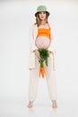 Young beautiful pregnant woman with long dark hair wearing green bucket hat standing, holding fresh bunch of carrots. Royalty Free Stock Photo