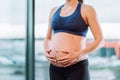 Unrecognisable pregnant woman in sportswear with chain and pendant on neck caressing belly on window background.