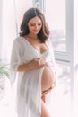 Young beautiful pregnant woman stand near window hugs bare belly with hands. White long silk negligee pregnancy dress Royalty Free Stock Photo