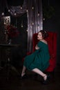 Young beautiful red-haired caucasian woman with professional makeup in green dress posing on red sofa Royalty Free Stock Photo