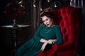 Young beautiful red-haired caucasian woman with professional makeup in green dress posing on red sofa Royalty Free Stock Photo