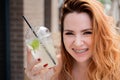 Young beautiful red-haired woman with braces drinks cooling lemonade outdoors in summer. Portrait of a smiling girl with Royalty Free Stock Photo