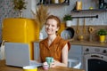 Young beautiful red-haired woman using a laptop computer and drinking a morning cup of coffee or tea. He looks at the camera and