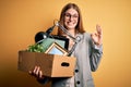 Young beautiful redhead businesswoman fired holding carboard box over yellow background doing ok sign with fingers, excellent
