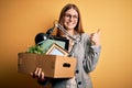 Young beautiful redhead businesswoman fired holding carboard box over yellow background happy with big smile doing ok sign, thumb