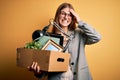 Young beautiful redhead businesswoman fired holding carboard box over yellow background stressed with hand on head, shocked with