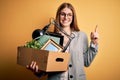 Young beautiful redhead businesswoman fired holding carboard box over yellow background surprised with an idea or question