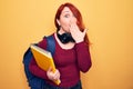 Young beautiful redhead student woman wearing backpack and headphones holding notebook covering mouth with hand, shocked and Royalty Free Stock Photo
