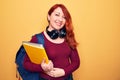 Young beautiful redhead student woman wearing backpack and headphones holding notebook looking positive and happy standing and Royalty Free Stock Photo