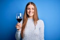 Young beautiful redhead woman drinking glass of red wine over  blue background with a happy face standing and smiling with Royalty Free Stock Photo