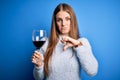 Young beautiful redhead woman drinking glass of red wine over isolated blue background with angry face, negative sign showing Royalty Free Stock Photo