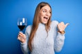 Young beautiful redhead woman drinking glass of red wine over isolated blue background pointing and showing with thumb up to the Royalty Free Stock Photo