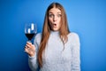 Young beautiful redhead woman drinking glass of red wine over isolated blue background scared in shock with a surprise face, Royalty Free Stock Photo