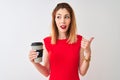 Young beautiful redhead woman drinking take away coffee over isolated white background pointing and showing with thumb up to the Royalty Free Stock Photo